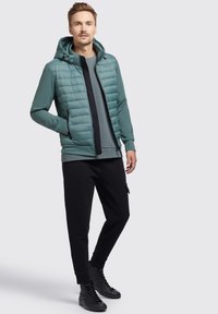 Ge puffte türkisfarbene Jacke mit Kapuze, schwarzem Frontreißverschluss, grauem Shirt darunter, kombiniert mit schwarzen Jogginghosen und schwarzen High-Top Sneakers.