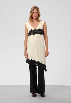 Femme portant une tunique crème sans manches avec bordure en dentelle noire sur un pantalon noir à jambes larges et des chaussures noires, debout devant un fond uni.