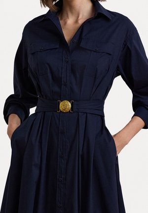 Mujer que lleva un vestido abotonado azul oscuro con las mangas remangadas y un cinturón a la cintura con una hebilla dorada con diseño de ancla.
