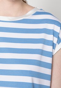 Camisa de rayas azules y blancas con tejido suave, escote amplio y ribete de satén en el cuello. Las mangas cortas tienen bordes enrollados.