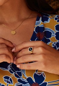 Zelts gredzens ar kvadrātveida melnu akmeni, nēsāts uz rokas. Ziedu raksta audums ar zilu, dzeltenu un sarkanu krāsu fonā.