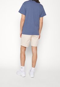Casual outfit featuring a blue cotton t-shirt and beige shorts. Footwear includes white sneakers with black accents. Plain background.
