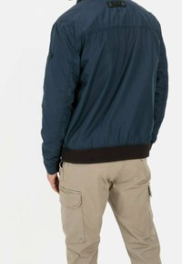 Granatowa kurtka bomberka o gładkiej fakturze, ze ściągaczem na dole i czarnym akcentem przy kołnierzu. Połączona z jasnobeżowymi spodniami typu cargo.