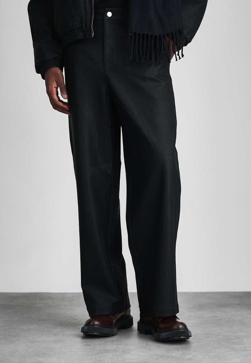 Pantalons en cuir noir avec une coupe décontractée, présentant une texture lisse et des coutures visibles, associés à des bottines marron à la cheville. Écharpe sombre à franges.