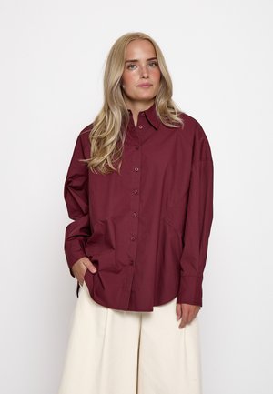 Giovane donna con lunghi capelli biondi indossa una camicia bordeaux larga con bottoni e pantaloni crema a gamba larga, in piedi davanti a uno sfondo bianco.