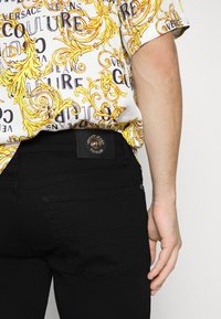 Vit skjorta med gult barockmönster och svarta jeans med en rund logotyp på en läderlapp nära midjan. Slät tygtextur.