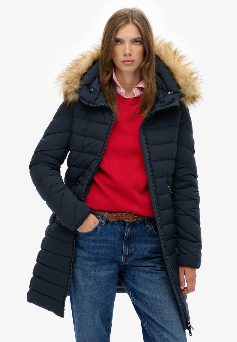 Mørkeblå pufferjakke med hætte med pelskant, har lynlåslukning, quiltet design og sidelommer, kombineret med en rød sweater og jeans.