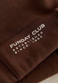 Brown fabric with white text reading "FUNDAY CLUB established since 1982" printed on a sleeve or cuff area.