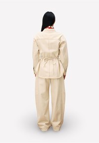 Person mit langen schwarzen Haaren steht mit dem Rücken zur Kamera, trägt eine beige Jacke, die an der Taille gebunden ist, und passende weit geschnittene Hosen vor einem weißen Hintergrund.