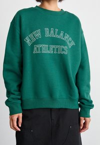 Sudadera verde de algodón con cuello redondo, con un estampado blanco curvado que dice “NEW BALANCE ATHLETICS”. Puños y dobladillo acanalados. Corte holgado.