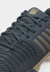 Σκούρο γκρι αθλητικό παπούτσι K-Swiss με γλώσσα από διχτυωτό ύφασμα, κορδόνια σταυρωτά και μπεζ διακριτικά λογότυπα στο πλάι και τη γλώσσα.