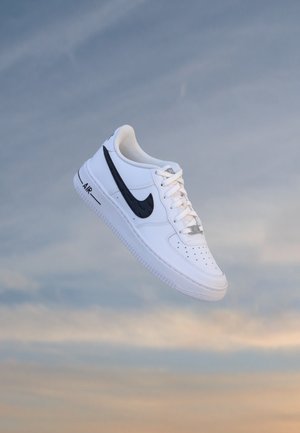 Άσπρο αθλητικό παπούτσι Nike Air Force 1 με μαύρο λογότυπο Swoosh που αιωρείται σε ένα απαλό ουρανό στο ηλιοβασίλεμα.