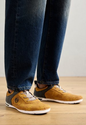 Yellow perforated sneakers with gray accents, featuring a textured surface, laces, and a white rubber sole, paired with dark denim pants.