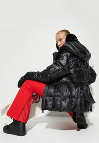 Chaqueta puffer negra con capucha, con un acabado brillante, diseño acolchado, cintura ajustada y detalles con cremallera, combinada con pantalones rojos y zapatillas negras.