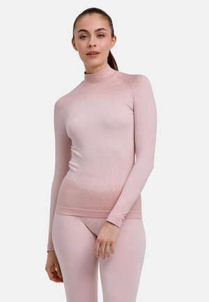 Frau mit braunen zurückgebundenen Haaren, die ein eng anliegendes hellrosa Langarmshirt und passende Leggings vor einem einfarbigen Hintergrund trägt.