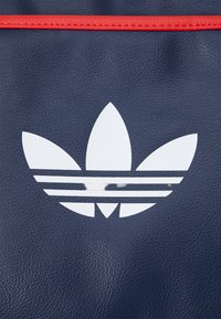 Saco de couro texturizado azul-marinho com um logo trefoil branco da Adidas acompanhado de três riscas horizontais por baixo, realçado por uma borda vermelha.