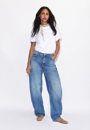 Donna con capelli lunghi e scuri che indossa una t-shirt bianca, una sciarpa chiara, jeans larghi blu e ballerine beige, in piedi davanti a uno sfondo bianco.