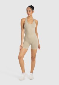 Body color beige con scollatura a barchetta e design a schiena scoperta. Texture liscia e forma aderente, abbinato a scarpe da ginnastica bianche.