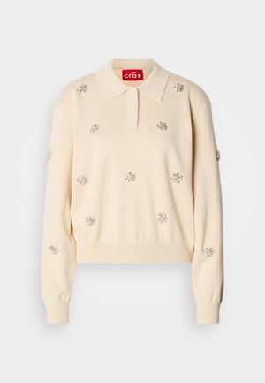 Pull en maille color crème, avec un col polo, orné de décorations florales en argent. Présente des poignets et un ourlet côtelés.