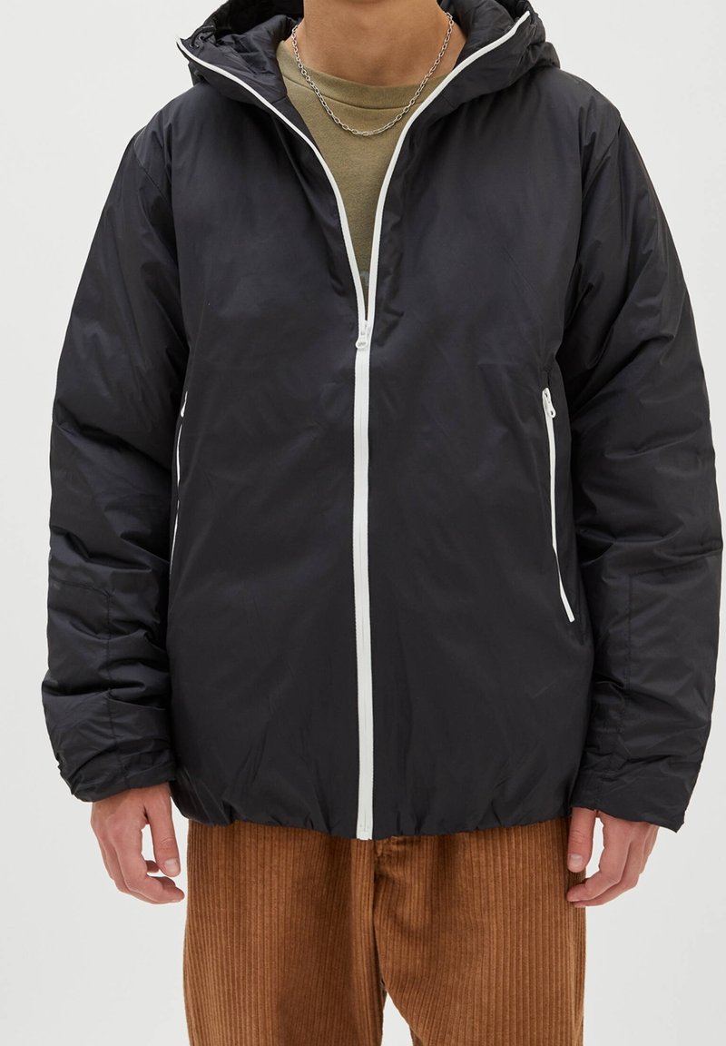 Veste matelassée noire avec capuche, fermeture éclair blanche, ourlet élastique et deux poches latérales. Matière lisse avec une finition mate.