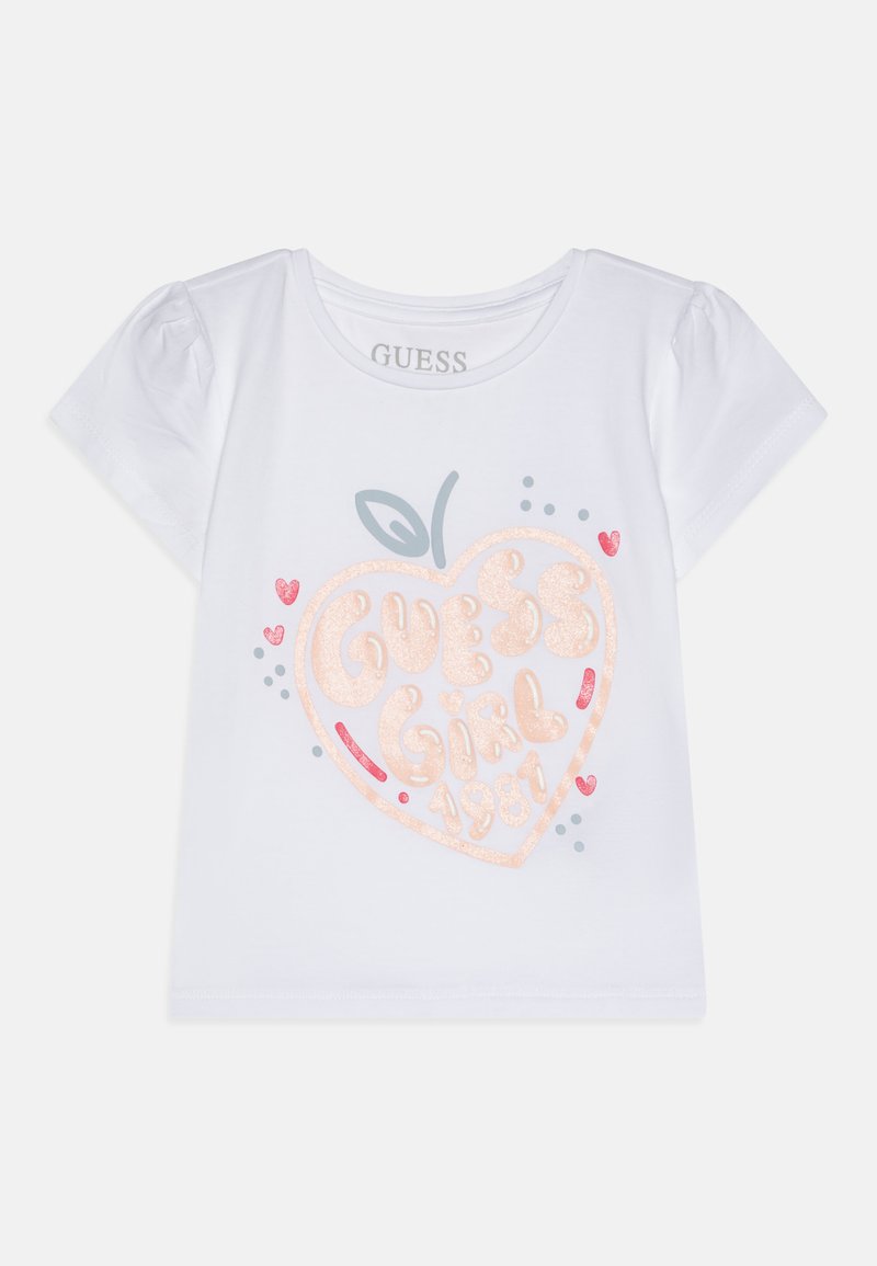 Hvit T-skjorte med korte ermer som har et rosa glitrende hjertegrafikk og teksten "GUESS Girl 1981", akkompagnert av små fargede prikker og hjerter.
