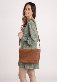 Brown leather shoulder bag with a smooth texture, featuring a detachable strap and a subtle logo patch. Model wears a green patterned dress.