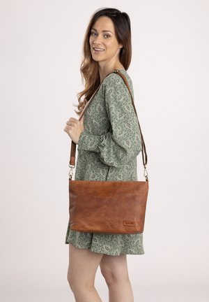 Borsa a spalla in pelle marrone con una texture liscia, dotata di una tracolla rimovibile e un discreto logo. Il modello indossa un vestito verde a fantasia.