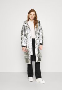 Casaco puffer refletivo prateado com capuz, fecho frontal e dois bolsos grandes. Usado sobre um suéter creme de gola alta e calças pretas, combinado com sapatilhas brancas.