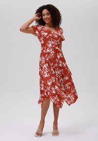 Robe florale rouge avec des motifs de roses blanches, décolleté en V, manches courtes, jupe de longueur genou avec un ourlet asymétrique, associée à des sandales blanches.