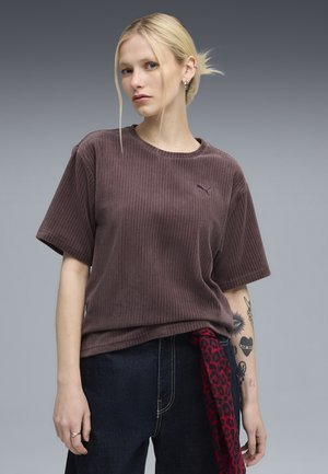 T-shirt marrone a costine con maniche corte e colletto rotondo; presenta un piccolo logo viola sul lato sinistro del petto. Il modello indossa pantaloni in denim scuro.