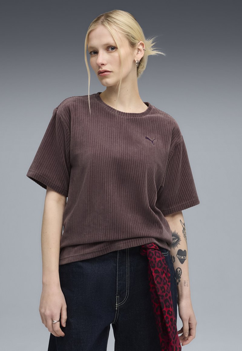 Brun ribbet T-shirt med korte ærmer og rund hals; har et lille lilla logo på venstre bryst. Modellen har mørke denimbukser på.