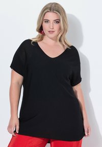 Černé volné tričko s výstřihem do V a krátkými rukávy. Měkká textura, uvolněný design a prodloužená zadní část, spojené s červenými texturovanými kalhotami.