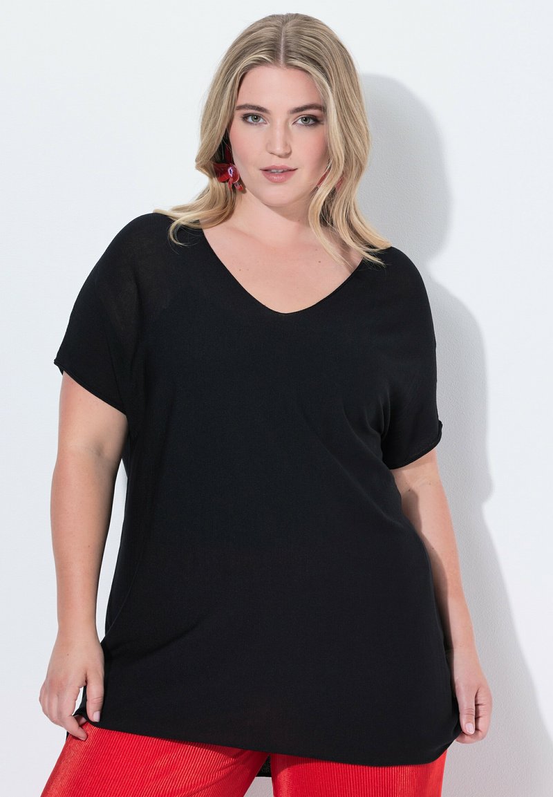 Černé volné tričko s výstřihem do V a krátkými rukávy. Měkká textura, uvolněný design a prodloužená zadní část, spojené s červenými texturovanými kalhotami.