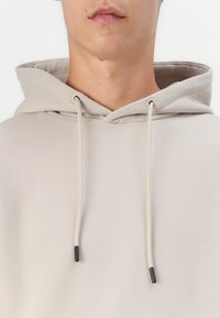 Beige Kapuzenpullover mit Kordelzügen; glatte Textur; elastische Kordelenden mit schwarzen Spitzen; einfaches Design, keine Muster oder Grafiken.