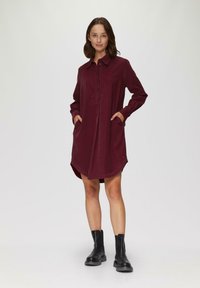 Weinrotes Hemdkleid mit knöpfbarem Kragen, langen Ärmeln, seitlichen Taschen und abgerundetem Saum. Die Stoffstruktur wirkt weich und glatt.