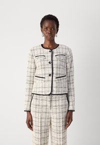 Veste en tweed crème avec des accents noirs, présentant un col rond, des boutons à l'avant et deux poches poitrine. Texture lisse avec un motif à carreaux.