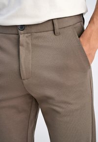 Pantaloni beige con una texture liscia, dotati di chiusura con bottone e tasche laterali. Il tessuto presenta un leggero motivo a intreccio diagonale.