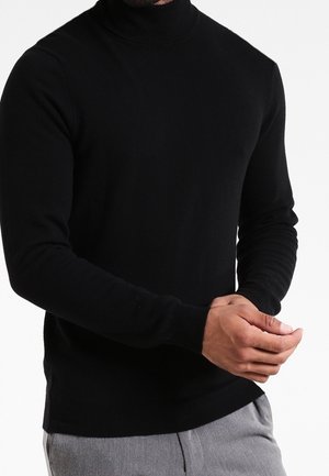 Homme portant un pull noir ajusté à col roulé et manches longues, ainsi qu'un pantalon gris clair, ajustant son poignet gauche avec sa main droite.