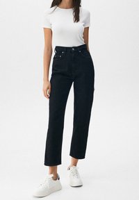 Jean en denim noir taille haute avec coupe droite, comprenant cinq poches, une braguette à boutons, et des baskets blanches.