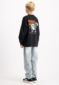 Zwart shirt met lange mouwen en een grafische print op de achterkant, gecombineerd met lichtblauwe wijde spijkerbroek en zwarte sneakers met witte strepen.