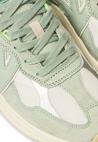 Sneaker verde chiaro caratterizzato da un mix di materiali in suede e rete, lacci prominenti e accenti bianchi a contrasto con dettagli testurizzati.