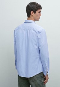Junger Mann, der seitlich steht und ein hellblaues langärmliges Hemd mit feinen Streifen sowie eine dunkle Jeans trägt, vor einem schlichten Hintergrund.