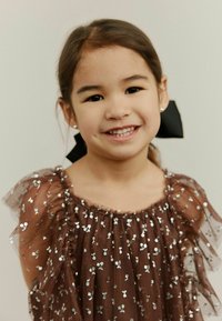 Barna tüllruha ezüst flitterekkel, fodros ujjal és ráncolt nyakkivágással. Fekete masnival feltűzött haj. Mosolygó gyermek.