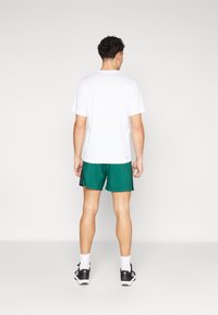 Weißes T-Shirt mit kurzen Ärmeln, kombiniert mit grünen Sportshorts aus atmungsaktivem Stoff, mit glatter Textur und normaler Passform. Schwarze Sneakers runden den Look ab.