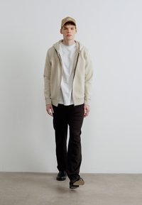 Beżowa bluza z kapturem na zamek na białej koszulce, zestawiona z czarnymi spodniami i czarnymi butami. Beżowa czapka dopełnia casualowy strój.