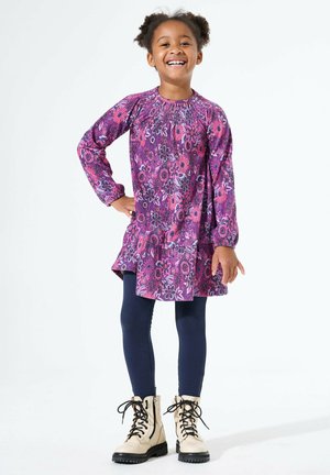 Mädchen lächelt mit einer Hand auf der Hüfte, trägt ein lila Blumenkleid, marineblaue Leggings und beige Schnürstiefel vor weißem Hintergrund.