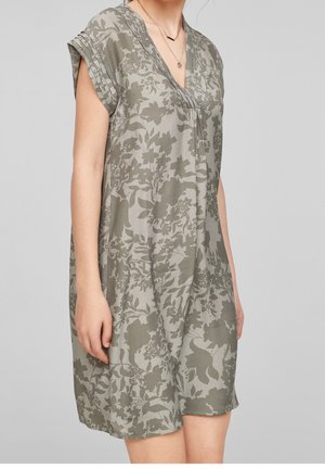 Femme portant une robe à manches courtes, longueur genoux, avec un motif floral abstrait vert et gris et un décolleté en V, montrée du cou aux genoux.