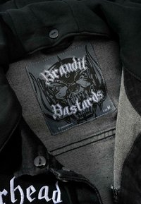 Svart tygjacka med knappstängning. Inneretikett med ett skallemönster och texten "Brandit Bastards," storlek M. Synlig sömnadsdetalj.