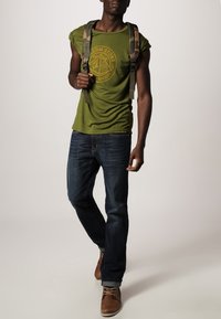 Olivegrøn kortærmet t-shirt med et cirkulært grafik, marineblå jeans og brune snørebåndssko. Rygsækken har et camouflage-mønster.
