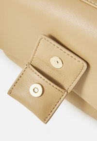 Beige leather bag detail featuring a snap closure with gold-tone hardware and contrasting stitching. Smooth texture and clean lines.